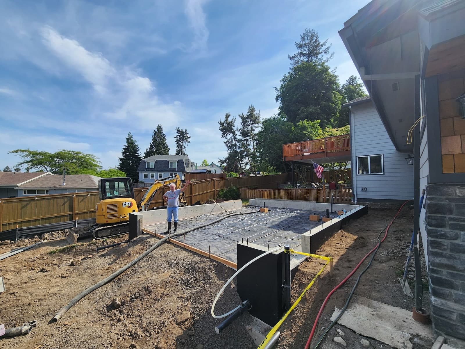 Poured concrete stem walls with anchor bolts set, a black vapor barrier laid over compacted base inside the forms, and a compact excavator parked alongside