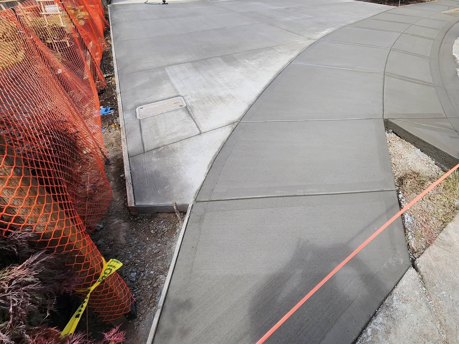 A newly poured driveway approach meeting a broad curved sidewalk run at a tooled joint, with a flush utility access lid set into the slab and orange safety fencing along the property line