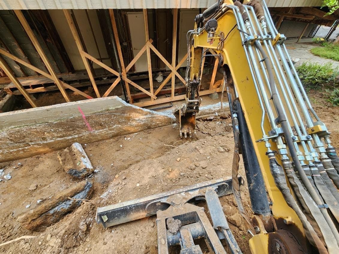An excavator arm reaching under a house held up on temporary timber cribbing, breaking out the old foundation below it, with a cut slab of the original concrete lying in the dug-out trench