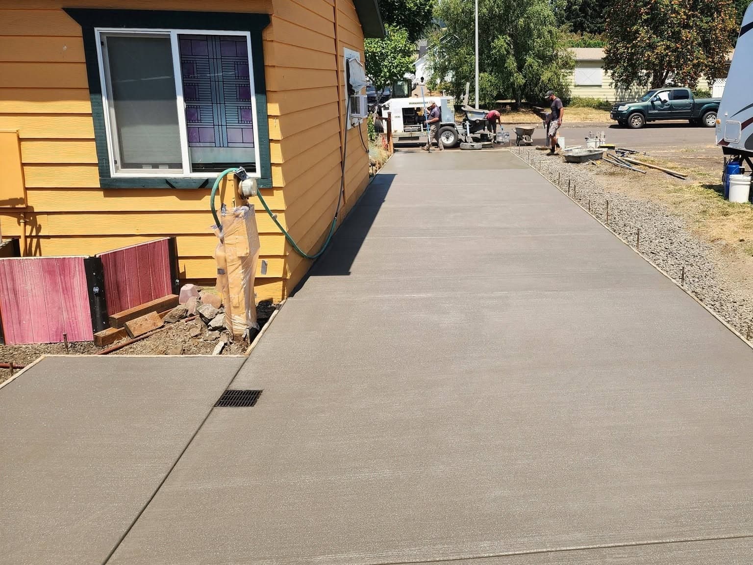 A long broom-finished concrete driveway running the length of a yellow house to the street, with a steel grated channel drain set flush into the slab in the near corner and fresh drain rock along the far edge
