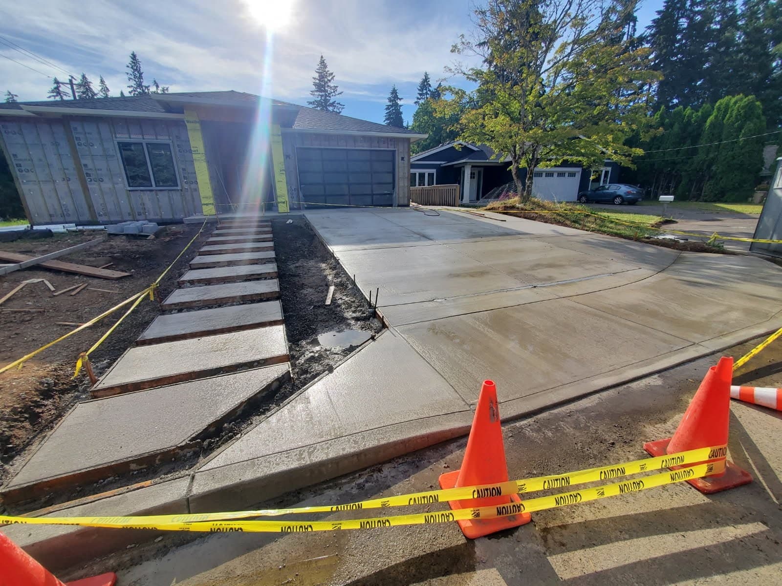 A complete new driveway still curing damp, running from a rounded street approach across a wide slab to the garage of a house under construction, with a stepped concrete walkway climbing alongside it and traffic cones and caution tape across the approach