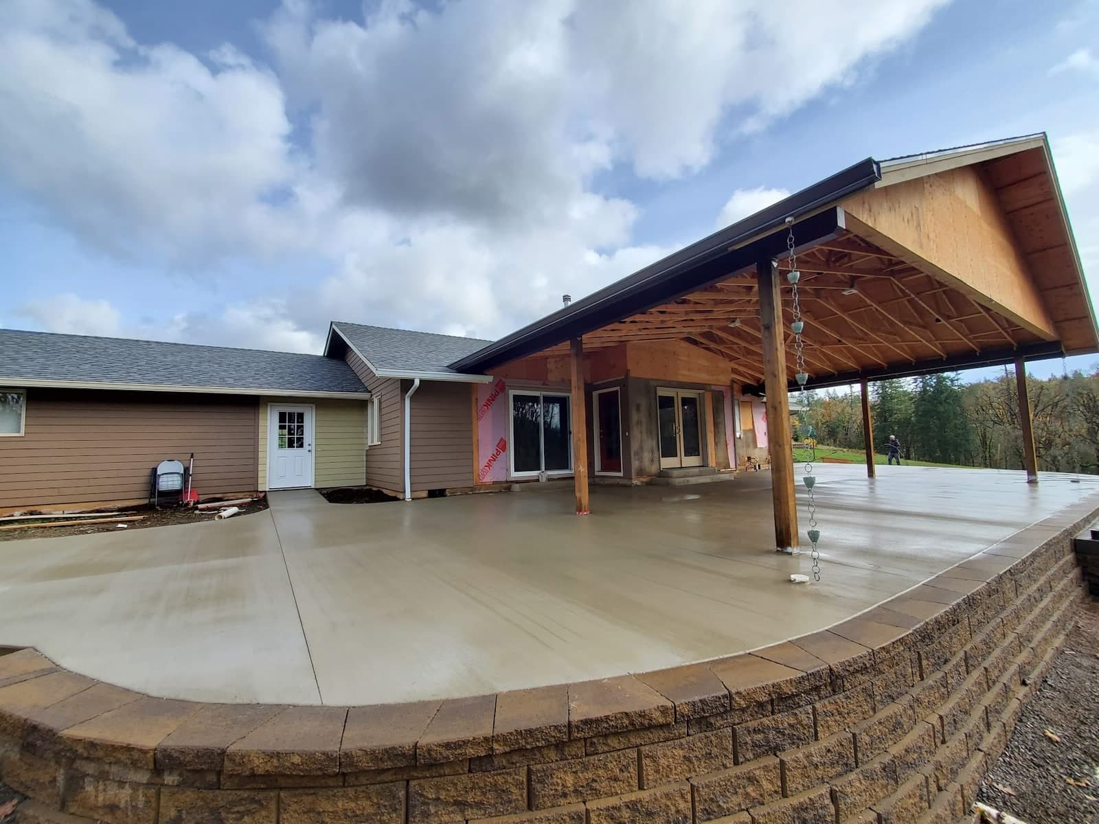 A wide, freshly poured concrete patio curving around the back of a house, still damp and broom-finished, running out under a newly framed covered porch with exposed rafters and timber posts