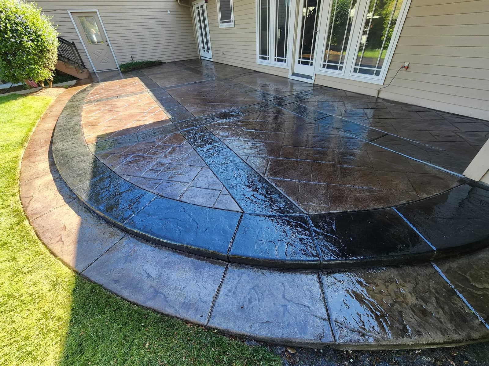 A restored stamped-concrete patio photographed close and low, the surface wet and deep red-brown again with a black banded ring sweeping across it, a tan outer border curving round the edge and a stamped step dropping to the lawn