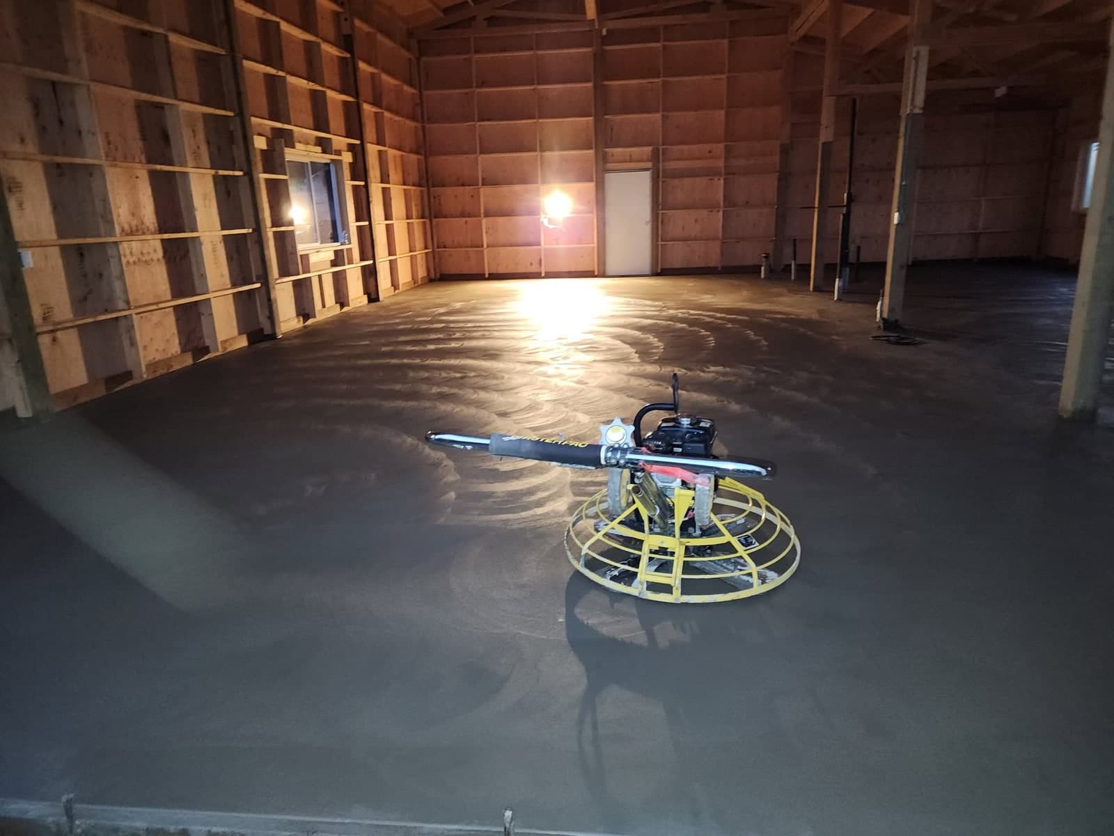 A broom-finished concrete slab and apron poured up to the wall of a dark steel shop building, with a yellow vapor barrier still laid out over the unpoured bay behind it and hills across the valley beyond