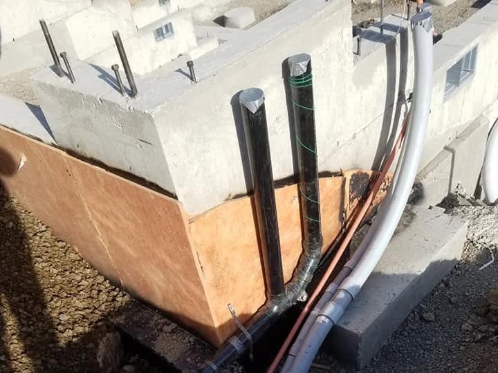 Gray PVC conduits and an orange flexible conduit crossing an open trench of wet clay, running past a poured concrete pier footing