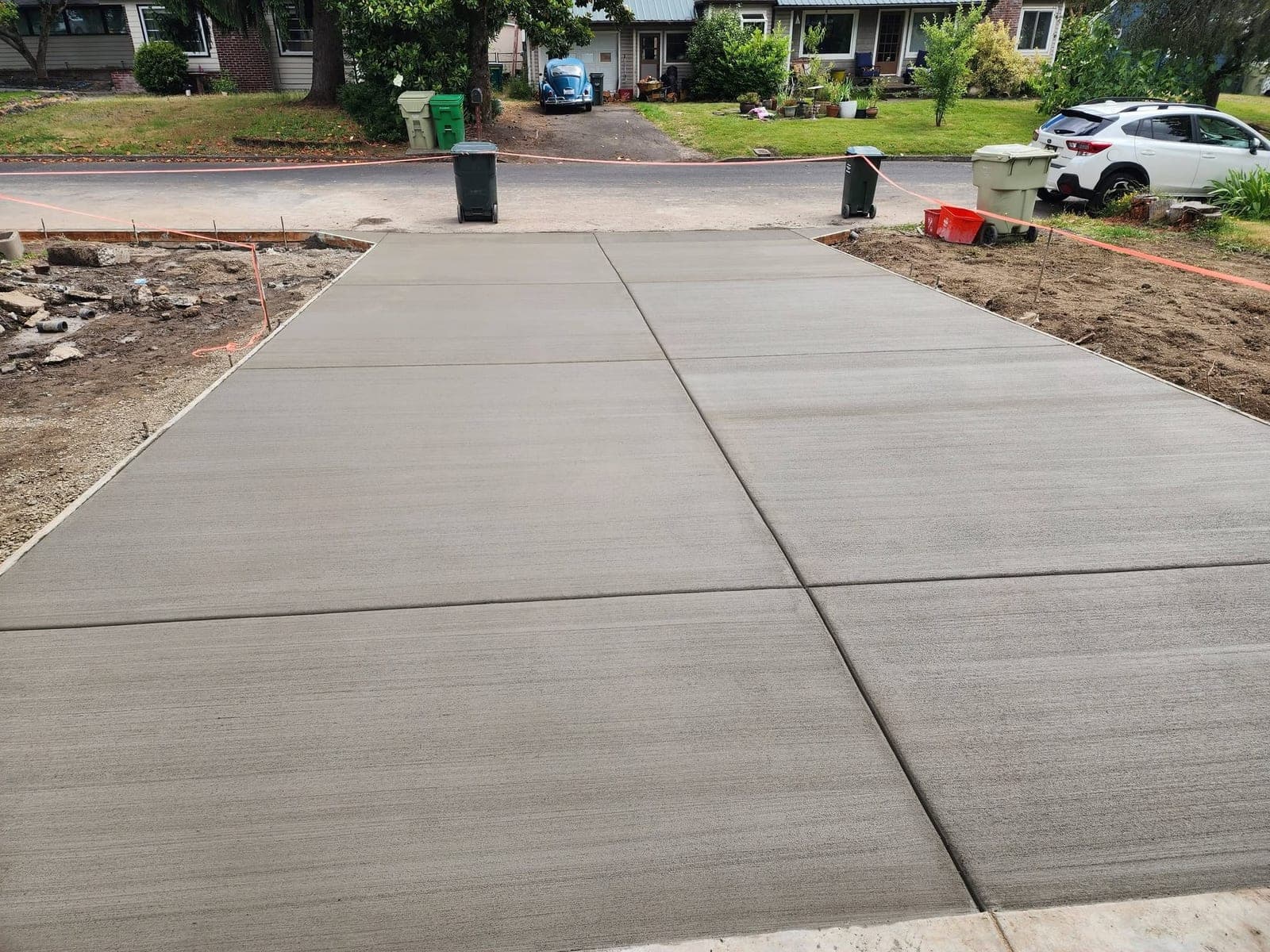 A wide new concrete driveway in Beaverton with a light broom finish and clean tooled control joints running from the foreground down to the street, freshly stripped soil either side and a residential street beyond
