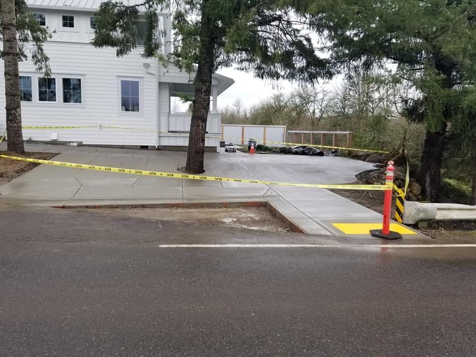 A finished concrete driveway in Carlton meeting the street through a flared approach, with a sidewalk crossing it and a rolled curb at the road edge, caution tape strung across the wet slab and a white farmhouse behind the firs