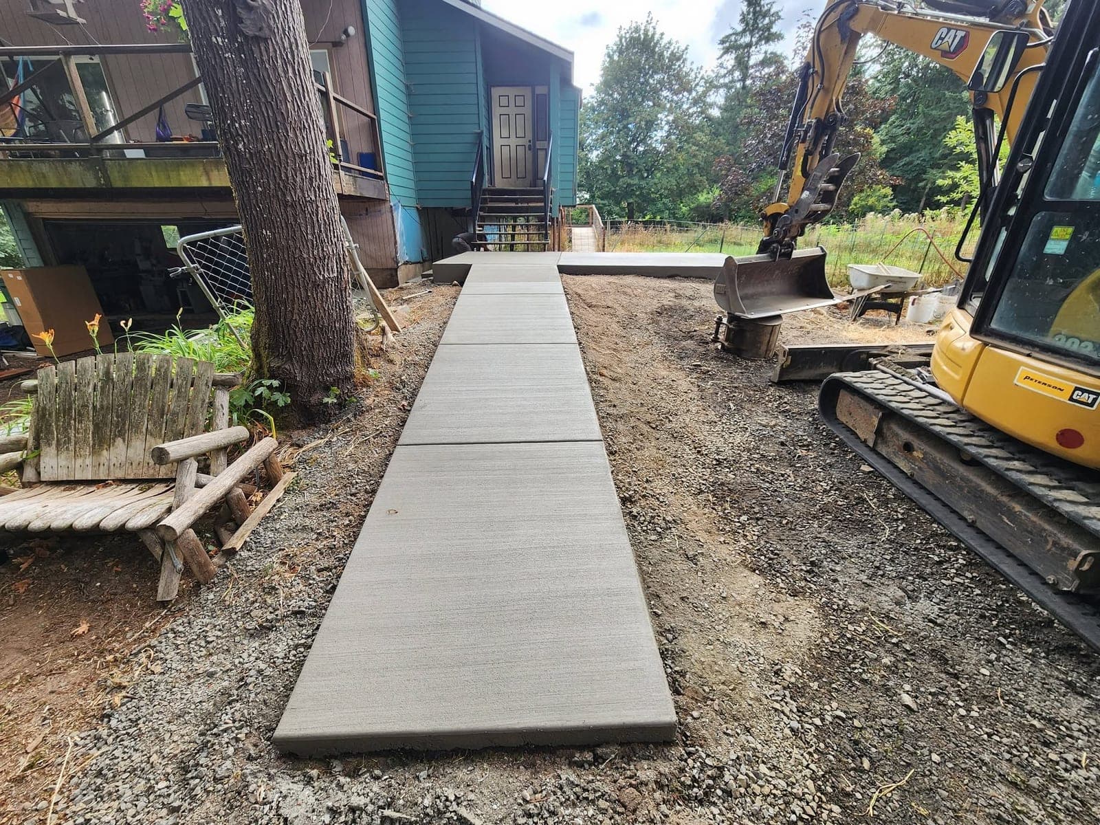 A broom-finished concrete walkway in Dundee running in three bays from a raised front entry out across bare graded dirt, with a tracked excavator parked alongside and a wheelbarrow beyond it
