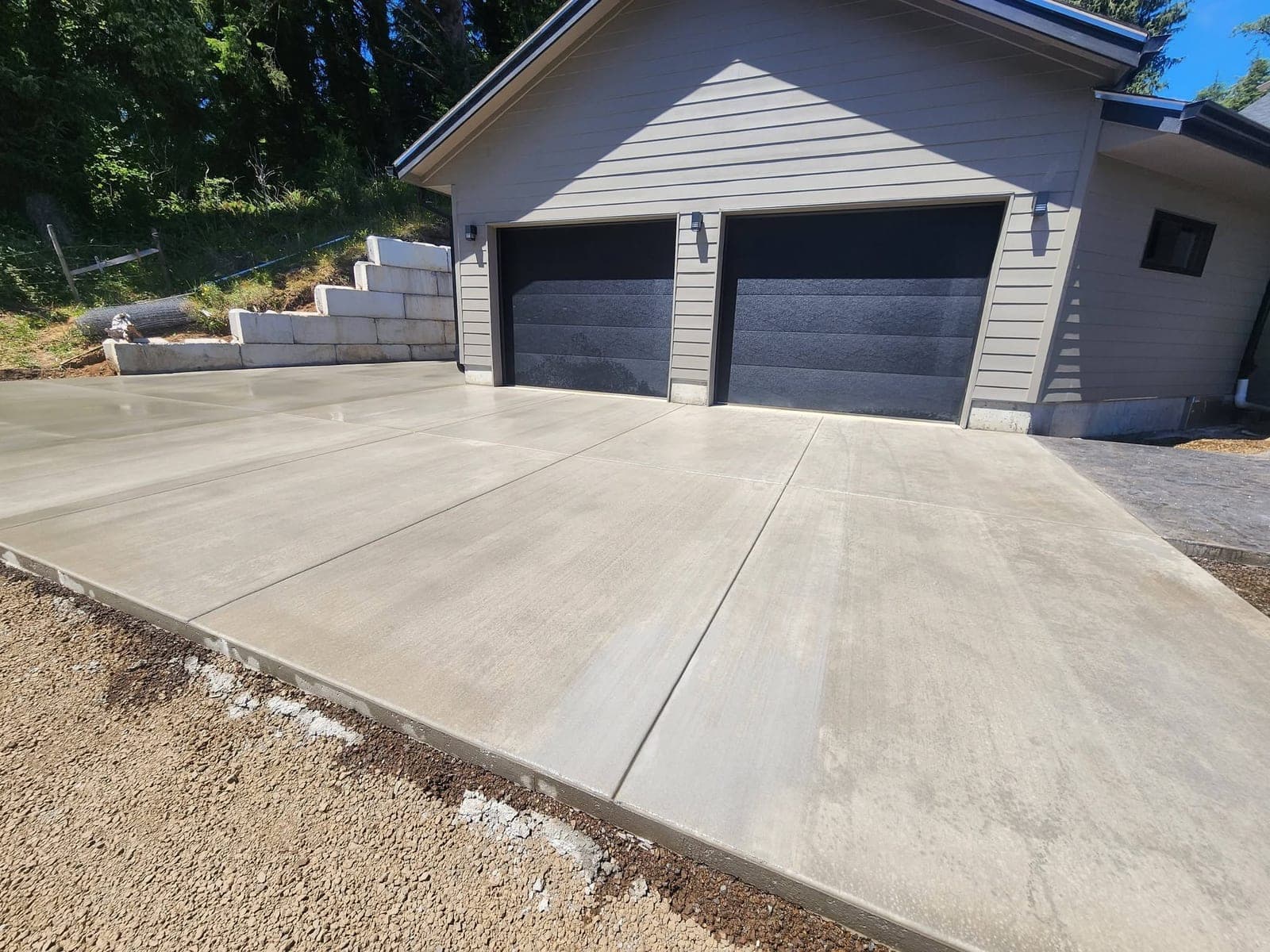 A wide broom-finished concrete driveway in Garibaldi running up to a double garage with tooled joints dividing it into bays, the gravel and dirt of the approach replaced edge to edge and a cut bank rising to firs on the left