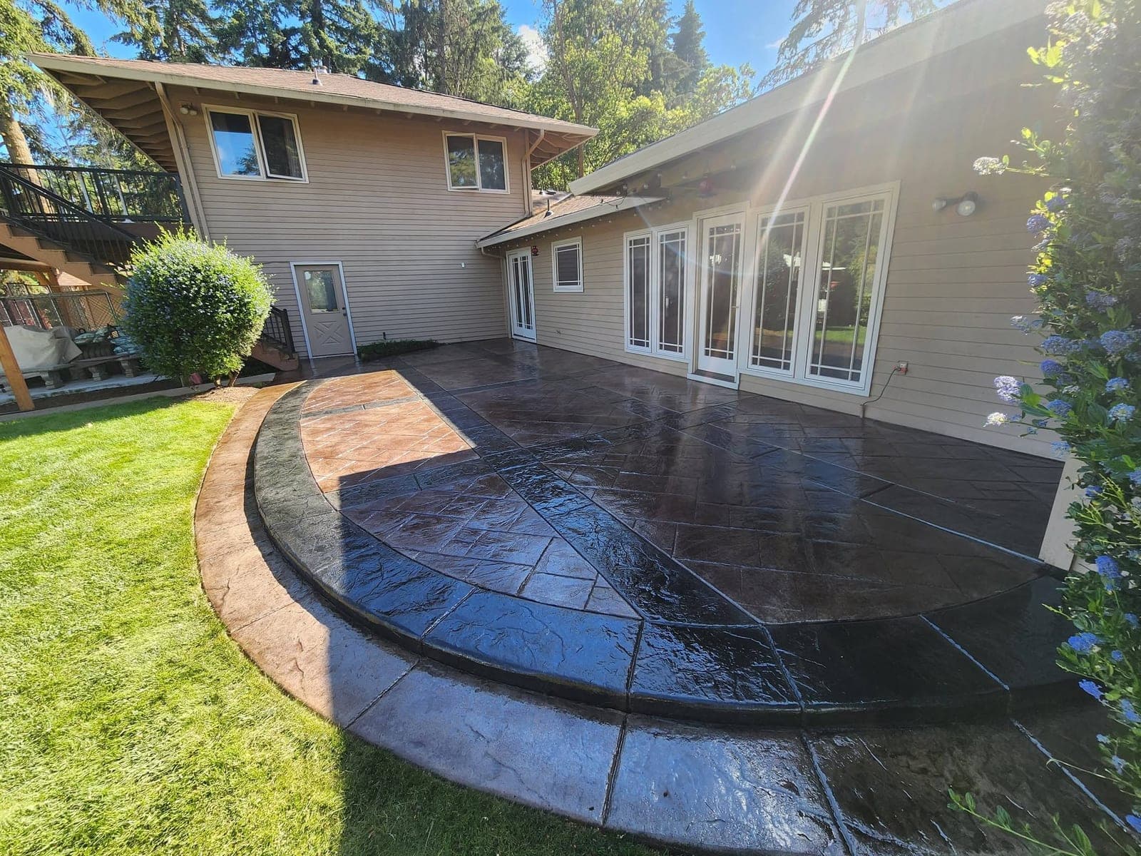 The same Hillsboro patio photographed wet, water lying across the ashlar pattern so the field and the bands read deep red-brown to near black and the house windows reflect in the surface, with one wedge of the outer border at the upper left still dry and light tan, curving round to a step down onto the lawn