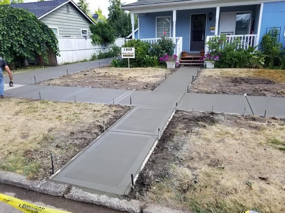 Freshly poured broom-finished concrete across the front of a pale blue McMinnville house: a walkway running from the street to the porch steps, a wide apron beside it and a further run along the property edge, with pins still standing in the wet edges