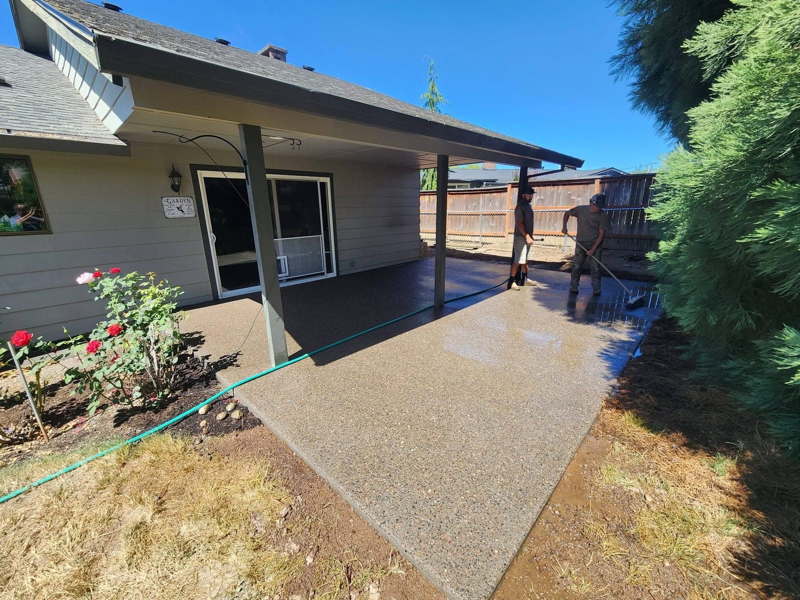 A large exposed-aggregate patio wrapping the back of a cream Newberg house under a covered porch, the near half dried to a pale pebbled finish and the far half still dark with rinse water, red roses in the foreground and two of the crew rinsing the far end