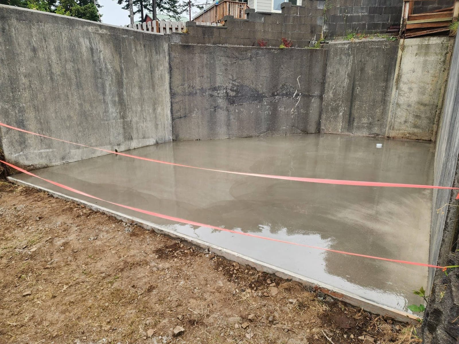 A smooth troweled concrete slab poured inside the existing walls of a dug-out foundation in Pacific City, still damp and sealed to an even sheen, with orange string lines strung across it at knee height