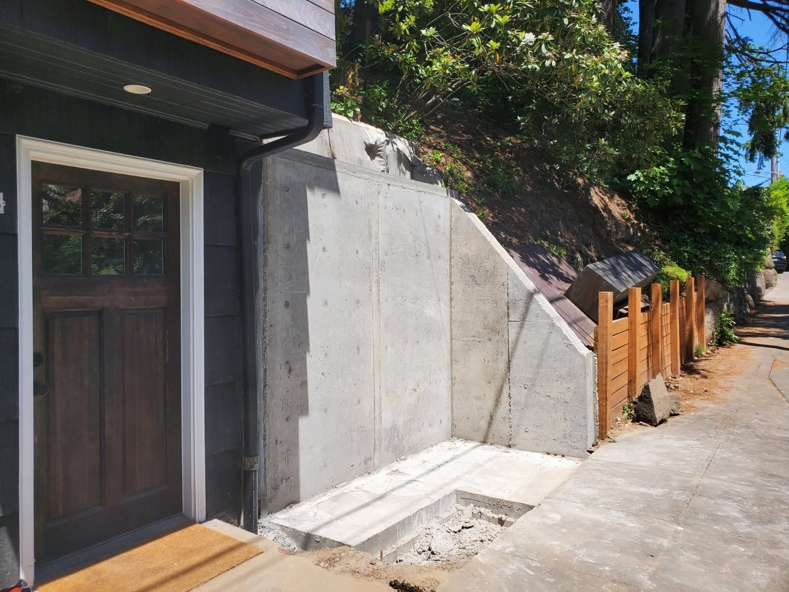 A board-formed concrete wall on a steep Portland street with its forms stripped, the gray face marked with tie-snap holes and its top stepping down to follow the grade, a poured pad at its foot and a cedar fence and pavement beyond