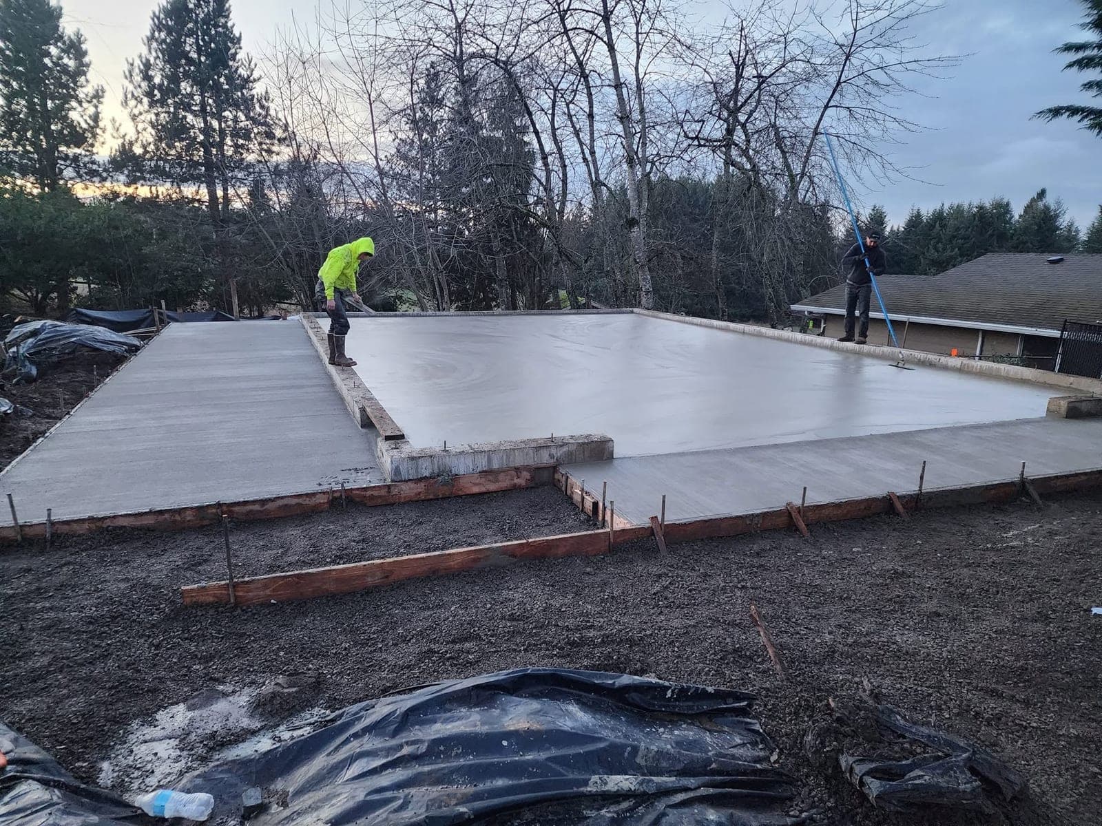A large freshly finished concrete slab filling the frame, still dark and damp, its perimeter held by timber forms with rebar dowels standing along the edge, one crew member in high-visibility yellow working the near edge and another drawing a broom across the far end