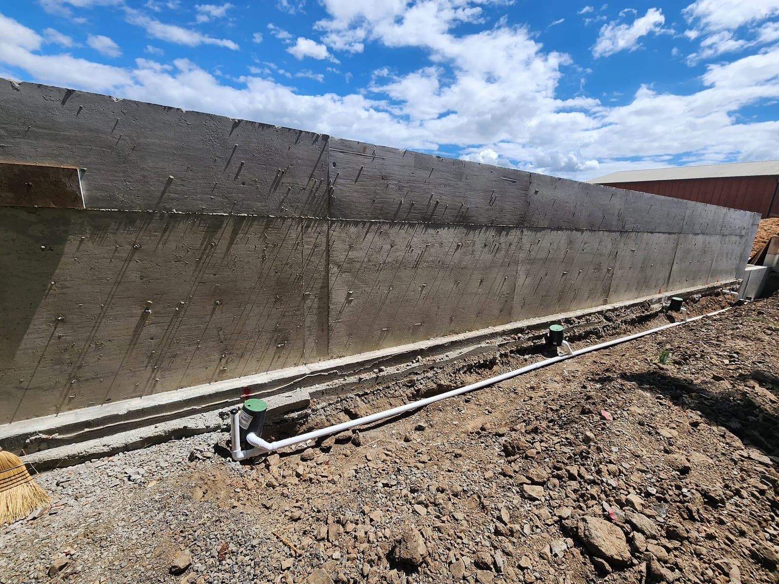 A long cast-in-place concrete wall running the width of the frame with the grain of the board forms still legible across its face and the tie holes in even rows, standing on a poured footing with a white drain line and green cleanout caps set along its base