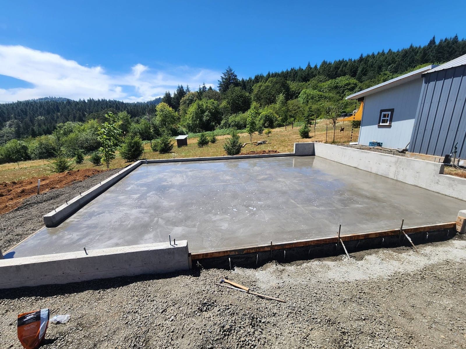 A large finished concrete slab in Yamhill poured inside low formed stem walls on a hillside pad, edged with crushed rock and anchor bolts, a blue steel shop building at the right and forested ridges beyond